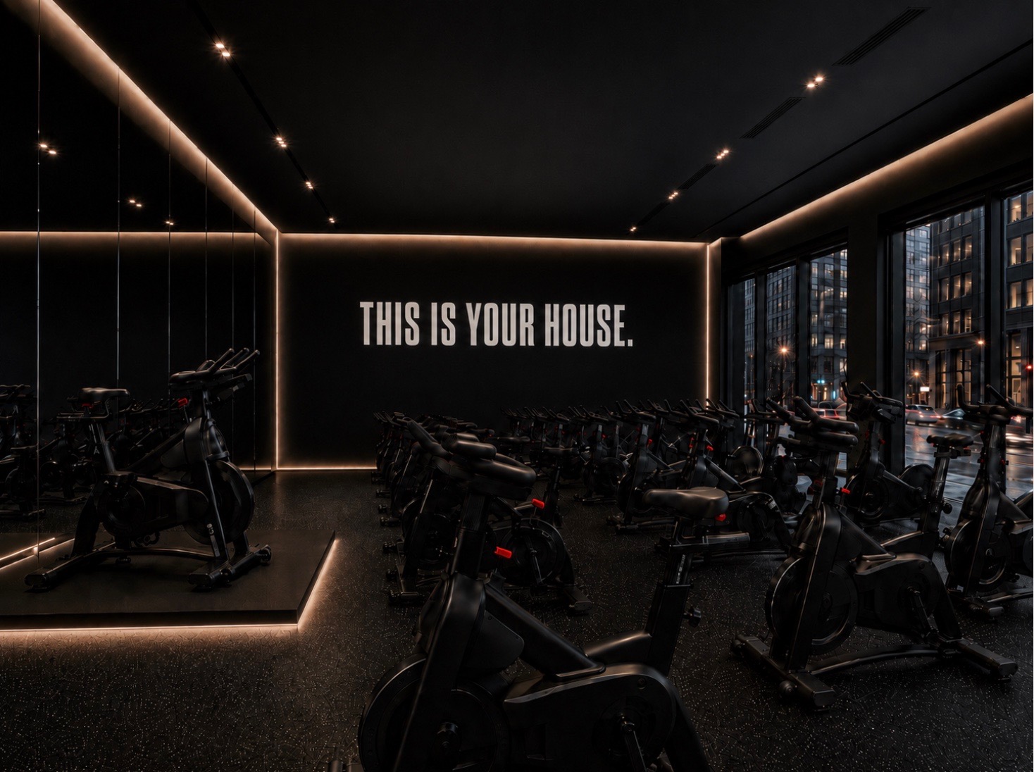 The Cycle House ride room: rows of black bikes under warm cove lighting, with 'This is your house.' on the wall
